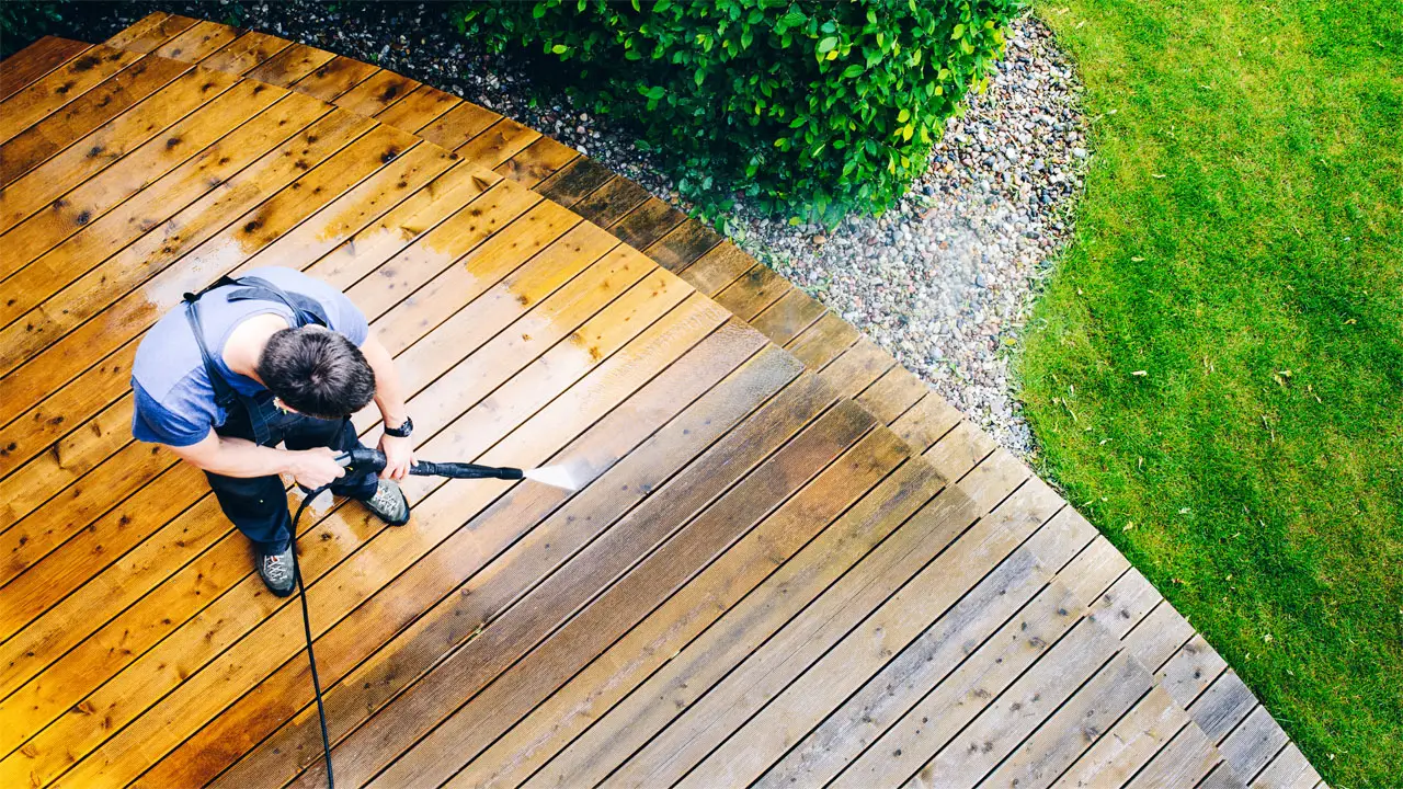 A man using a pressure washer to clean a wooden deck, spraying water to remove dirt and debris.